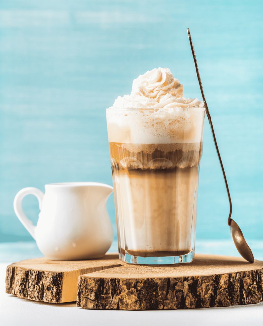 A glass of coffee with whipped cream and spoon on wooden table.